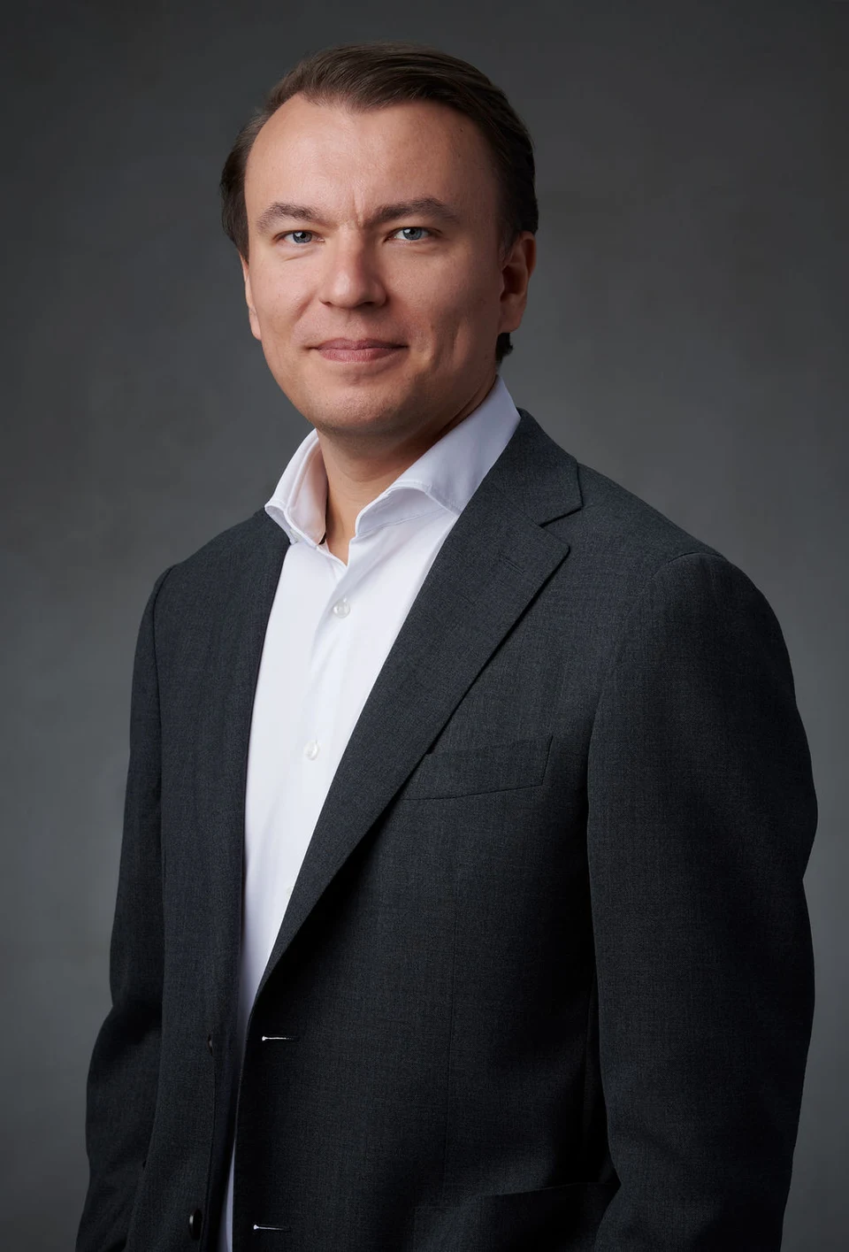 Stan Sabitovs in a dark suit and open-collar white shirt, standing against a neutral studio background.